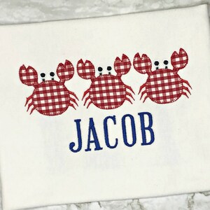 K&ouml;nnte beinhalten: Wei&szlig;es T-Shirt mit drei roten und wei&szlig;en Gingham-Krabben und dem Namen "JACOB" in Blau gestickt.