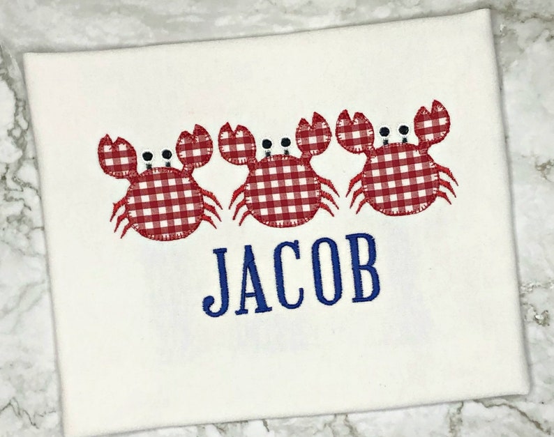 K&ouml;nnte beinhalten: Wei&szlig;es T-Shirt mit drei roten und wei&szlig;en Gingham-Krabben und dem Namen "JACOB" in Blau gestickt.