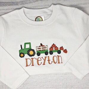 May include: White long-sleeve shirt with an embroidered design featuring a green tractor pulling a wagon filled with pumpkins. The name "Dreyton" is embroidered in orange script below the wagon. A fall-themed children's shirt.