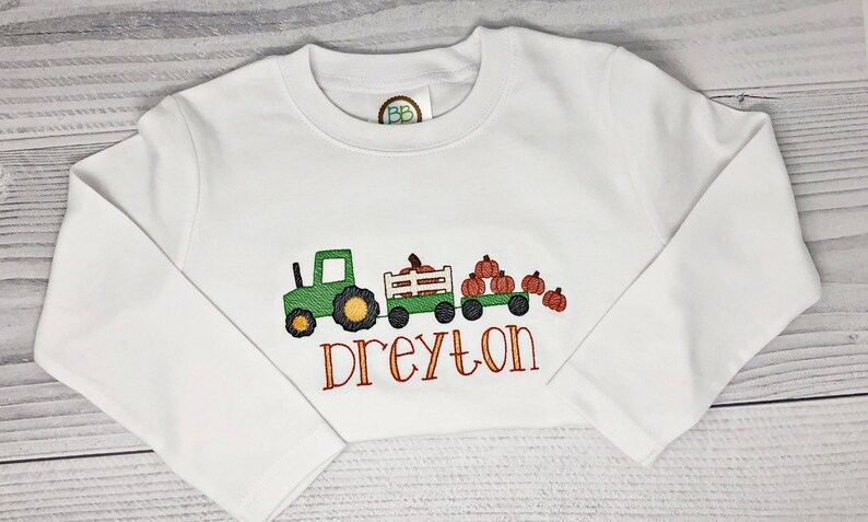 May include: White long-sleeve shirt with an embroidered design featuring a green tractor pulling a wagon filled with pumpkins. The name "Dreyton" is embroidered in orange script below the wagon. A fall-themed children's shirt.