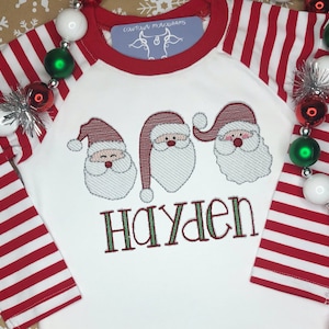 May include: White long-sleeved shirt with red and white striped sleeves. The shirt features three embroidered Santa faces and the name "Hayden" in green.