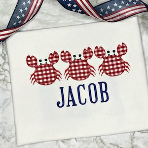 K&ouml;nnte beinhalten: Wei&szlig;es T-Shirt mit drei rot-wei&szlig; karierten Krabben und dem Namen "JACOB" in blau gestickt.