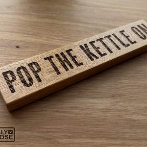 May include: A rectangular wooden sign with the phrase "POP THE KETTLE ON" etched in black. The sign is made of light-coloured wood and has a natural wood grain. The Jelly Moose logo is in the bottom left corner.