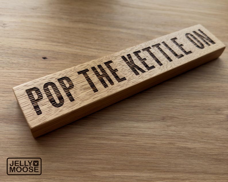 May include: A rectangular wooden sign with the phrase "POP THE KETTLE ON" etched in black. The sign is made of light-coloured wood and has a natural wood grain. The Jelly Moose logo is in the bottom left corner.