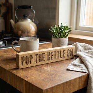 May include: A rectangular wooden sign with the words "POP THE KETTLE ON" engraved. It sits on a wooden surface, alongside a mug, a teapot, and a small potted plant. A linen cloth is also visible. The scene evokes a cozy kitchen setting.