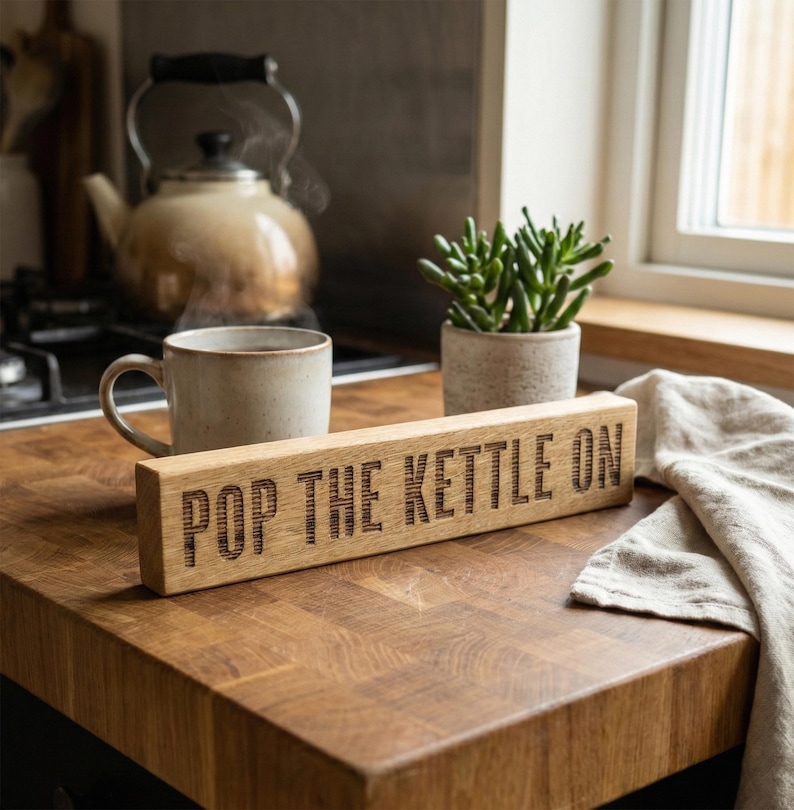 May include: A rectangular wooden sign with the words "POP THE KETTLE ON" engraved. It sits on a wooden surface, alongside a mug, a teapot, and a small potted plant. A linen cloth is also visible. The scene evokes a cozy kitchen setting.