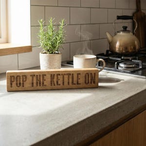 May include: A rectangular wooden sign with the words "POP THE KETTLE ON" engraved on it. The sign is on a kitchen counter, next to a plant and a cup of coffee. A kettle sits on the hob in the background. The countertop is made of concrete.