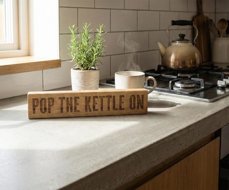 May include: A rectangular wooden sign with the words "POP THE KETTLE ON" engraved on it. The sign is on a kitchen counter, next to a plant and a cup of coffee. A kettle sits on the hob in the background. The countertop is made of concrete.