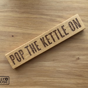 May include: A rectangular wooden sign with the phrase "POP THE KETTLE ON" engraved in dark letters. The sign is made of light-coloured wood and rests on a wooden surface. The Jelly Moose logo is in the bottom left corner.