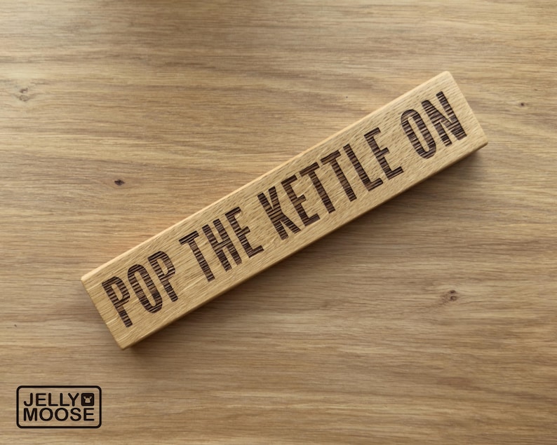 May include: A rectangular wooden sign with the phrase "POP THE KETTLE ON" engraved in dark letters. The sign is made of light-coloured wood and rests on a wooden surface. The Jelly Moose logo is in the bottom left corner.