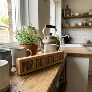 May include: A kitchen interior with a wooden sign reading "POP THE KETTLE ON". The sign is on a wooden shelf with mugs, a vintage teapot, and a potted plant. The kitchen has a window and a white tiled backsplash.