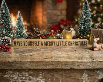 Könnte beinhalten: Ein rustikales Holzschild mit dem Schriftzug "HAVE YOURSELF A MERRY LITTLE CHRISTMAS" ist von Miniatur-Weihnachtsbäumen, Tannenzapfen und Lichterketten umgeben. Eine kleine Rentierfigur und Geschenkboxen vervollständigen die Weihnachtsszene.