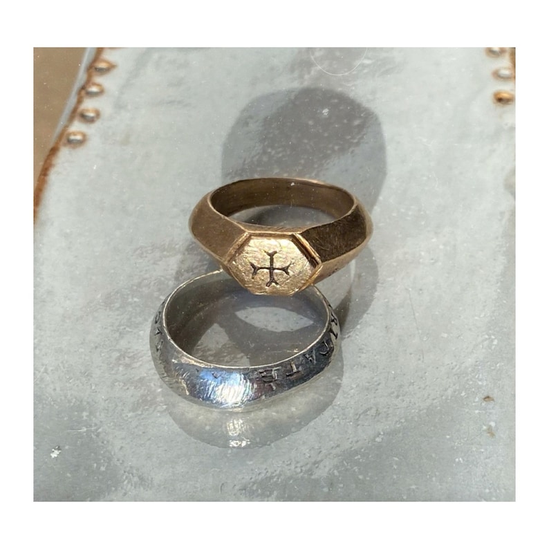 May include: Two rings, one gold and one silver, are stacked on top of each other. The gold ring has a hexagonal top with a cross design. The silver ring has a smooth, rounded surface.