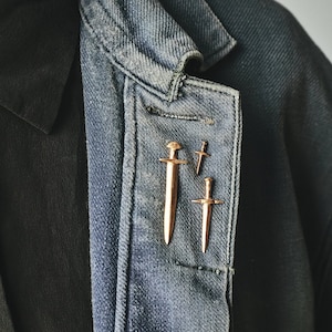 May include: Three gold-toned sword-shaped pins are pinned to a denim jacket. The pins are different sizes, with the largest pin in the center.