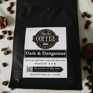 May include: A black bag of Fight Club Coffee labeled "Dark & Dangerous" with a description of "Dark Cocoa, Light Smoke, Syrupy, Ripe Black Cherry" and a roast level indicator. The bag is surrounded by coffee beans.