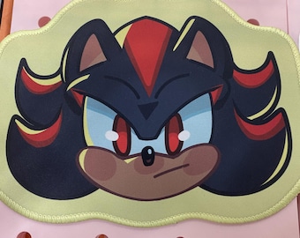 May include: A yellow mousepad featuring a cartoon image of Shadow the Hedgehog. The character has black fur with red highlights, red eyes, and a stern expression. The mousepad has a light yellow border and a pink background.