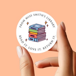 Peut inclure: Un autocollant rond avec une pile de livres colorés et le texte "From Miss Smith's Library. Read it. Love it. Return it." L'autocollant est tenu par une main sur un fond pêche.