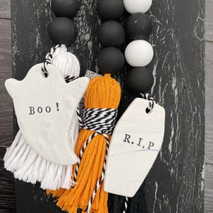 May include: Two Halloween door hangers with black and white beads. One hanger has a white ghost with the word "BOO!" and a white tassel. The other hanger has a white coffin with the text "R.I.P." and an orange tassel.