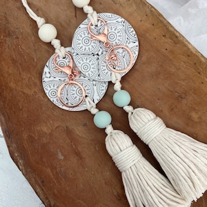 May include: A white cord with two white and blue beads and two white ceramic round keychains with black geometric designs and copper key rings. The cord has two white tassels attached.