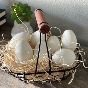 May include: A black wire basket filled with white eggs and brown wooden handles. The basket is sitting on a light brown surface with a green plant in the background.