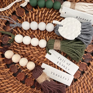 May include: A collection of decorative beads, tassels, and tags arranged on a woven basket. The beads are in shades of green, white, and natural wood. The tags read "Blessed", "full of thanks", and "Thankful".