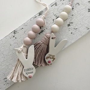 May include: Two white ceramic bunny charms with tassels. One charm says "Bunny Kisses" and the other says "Hello Spring". Both charms have pink accents and are strung on a beaded chain.