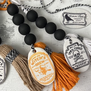 May include: A Halloween garland featuring black beads, orange and brown tassels, and white ceramic tags with black lettering. The tags read "Witch's Brew", "Crows Foot", "Unfiltered", and "Hemlock Root".