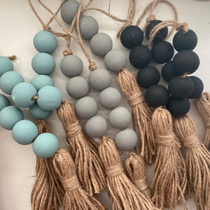 Peut inclure: Un ensemble de trois guirlandes de perles avec des glands. Les guirlandes sont faites de perles en bois bleu clair, gris et noir. Les glands sont en jute naturel.