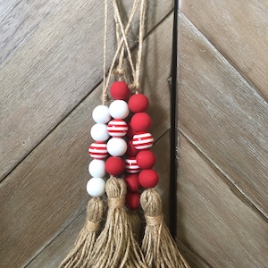 May include: A red and white beaded garland with a burlap tassel. The garland is made of wooden beads and is strung on a twine cord.