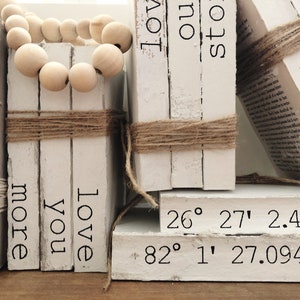 May include: A stack of white books with the words "home", "more", "you", "love", "love", "our", "story", and "blessed" printed on them. The books are tied with twine and decorated with a wooden bead garland. The bottom book has the coordinates "26° 27' 2.4804" and "82° 1' 27.0948" printed on it.