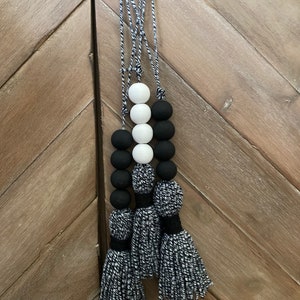 Puede incluir: Un tope de puerta de cuentas negras y blancas con tres borlas negras y blancas. El tope de puerta está colgado de un gancho en una pared de madera.