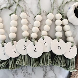 Puede incluir: Siete guirnaldas de cuentas de madera blancas con borlas verdes, cada una con una etiqueta redonda blanca numerada del 1 al 7.