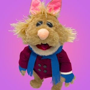 May include: A plush rabbit puppet with shaggy tan fur, large black eyes, and a pink nose. It wears a deep red double-breasted jacket with dark buttons, a bright blue scarf, and matching blue fingerless gloves. The mouth is open, revealing a heart.