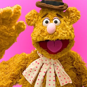 May include: A fuzzy golden-brown bear puppet with a brown fedora hat, a pink nose, and a purple tongue. The bear wears a white necktie featuring a pattern of small pink polka dots and is captured in a waving pose against a bright pink background.