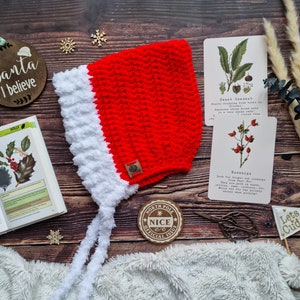 May include: A red and white crocheted Santa hat with a white pom-pom on the end of the tie. The hat is on a wooden surface with a book, a small wooden sign that says "Santa I believe", and a small wooden sign that says "North Pole Nice 10/10 Official Issue".