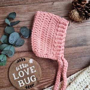 May include: A pink crocheted baby bonnet with a long tie and a tassel. The bonnet is lying on a wooden surface with a sign that says "little love bug".