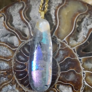 May include: A clear iridescent glass pendant with a gold chain. The pendant has a teardrop shape and is set against a brown background.