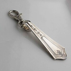 Puede incluir: Un llavero de plata hecho con un mango de cubiertos vintage. El mango tiene un diseño ornamentado y está unido a un cierre giratorio de plata.