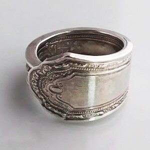 Può includere: Un anello d'argento realizzato da un cucchiaio riutilizzato. L'anello presenta un design decorativo e una finitura liscia e lucida.