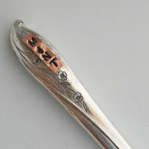 Puede incluir: Un cuchillo de mantequilla de plata con un diseño floral en el mango. El mango tiene una etiqueta de cobre con la inscripción "M22".