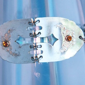 May include: A silver bracelet with a cross-shaped design and two orange gemstones. The bracelet is made from repurposed silverware.
