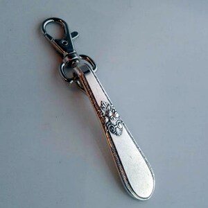 Puede incluir: Un llavero de plata con un diseño decorativo y adornado. La cadena tiene un clip de plata.