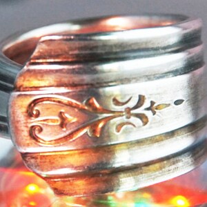 May include: A silver ring made from a repurposed spoon with a decorative design on the front. The ring is shiny and has a slightly worn look.