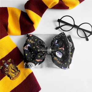 May include: A black and white Harry Potter themed bow with a gray headband. The bow features a repeating pattern of Harry Potter glasses, the number 9 3/4, a golden snitch, and a wizard's hat. The bow is tied with a silver ribbon.