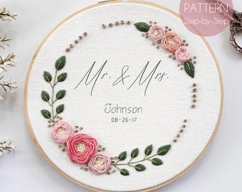 May include: Embroidered hoop art with a floral wreath design. The wreath is made of pink flowers and green leaves. The text "Mr. & Mrs. Johnson 08-26-17" is embroidered in the center of the hoop.