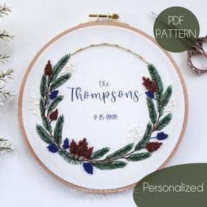 May include: A personalized embroidery pattern featuring a wreath of pine branches, berries, and pine cones. The text "the Thompsons 2-15-2020" is embroidered in the center of the wreath.