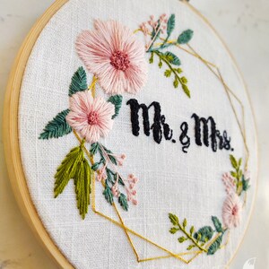May include: Embroidered hoop art with a gold geometric frame, pink flowers, green leaves, and the text "Mr. & Mrs."