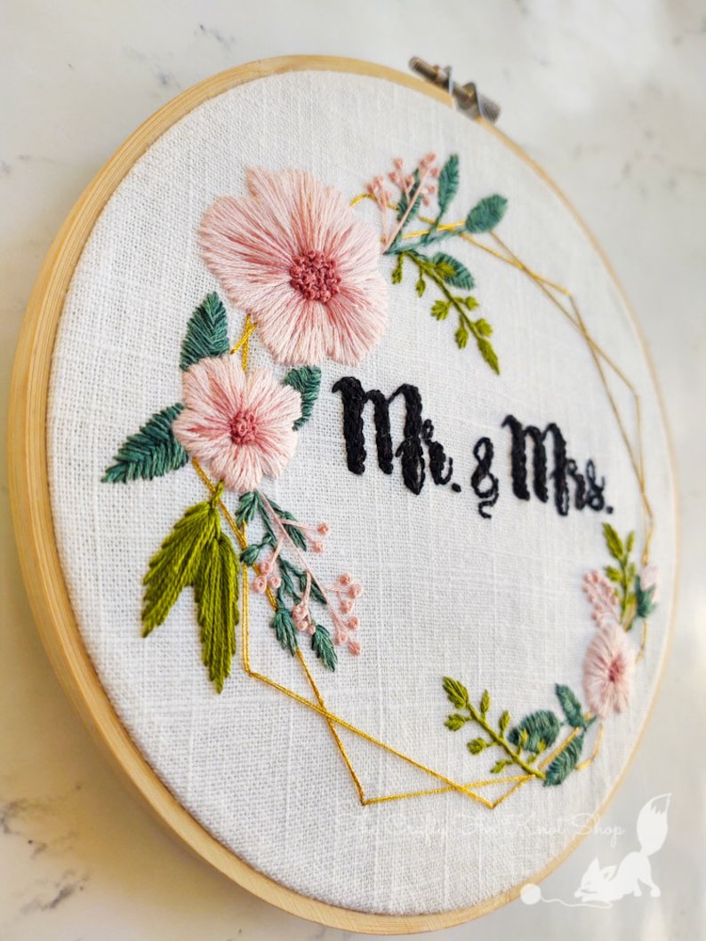 May include: Embroidered hoop art with a gold geometric frame, pink flowers, green leaves, and the text "Mr. & Mrs."