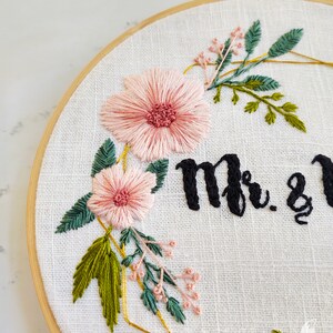 May include: Embroidered hoop art with pink flowers, green leaves, and gold geometric shapes. The text "Mr. & Mrs." is stitched in black thread.