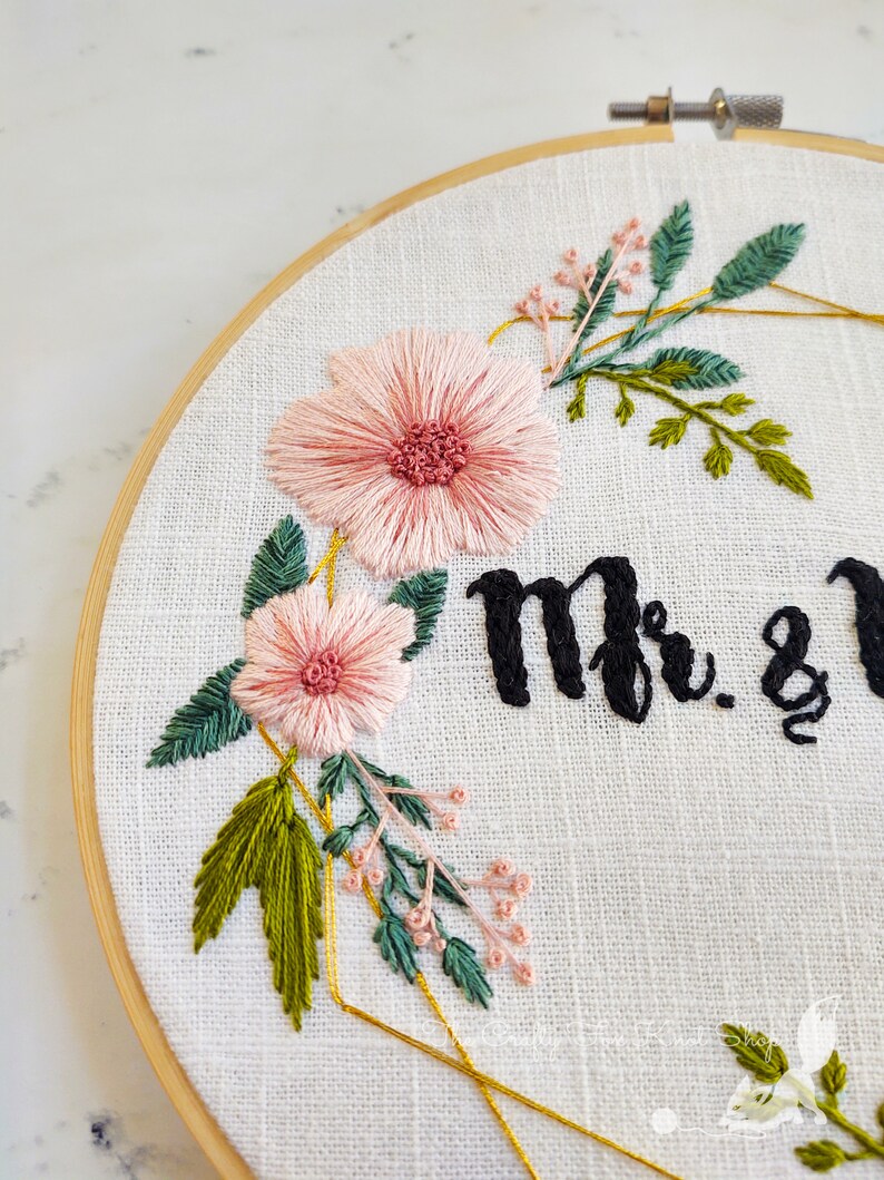 May include: Embroidered hoop art with pink flowers, green leaves, and gold geometric shapes. The text "Mr. & Mrs." is stitched in black thread.